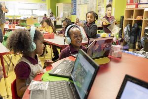 Two children smile as they complete their EarlyBird assessment on a chromebook, surrounded by a classroom of other students in instructional groupings